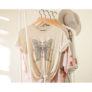 May include: A beige t-shirt with a black and white mandala butterfly design. The shirt is hanging on a white rack with other clothing.
