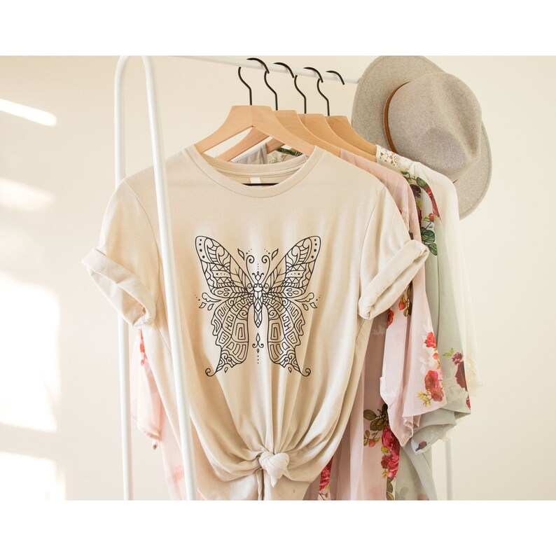 May include: A beige t-shirt with a black and white mandala butterfly design. The shirt is hanging on a white rack with other clothing.