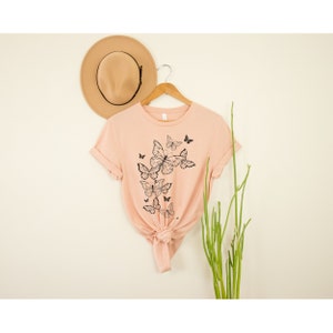 May include: A light pink t-shirt with a black butterfly graphic print. The shirt has a crew neck and short sleeves.