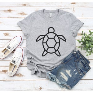Puede incluir: Una camiseta gris claro con un contorno negro de tortuga. La camiseta se combina con pantalones cortos de mezclilla y zapatillas de lona blancas. Los art&iacute;culos est&aacute;n colocados sobre una superficie de madera blanca con una planta verde.