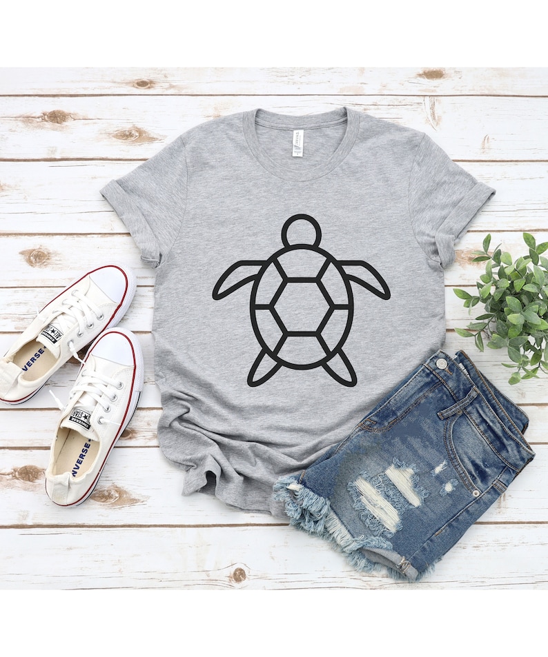 Puede incluir: Una camiseta gris claro con un contorno negro de tortuga. La camiseta se combina con pantalones cortos de mezclilla y zapatillas de lona blancas. Los art&iacute;culos est&aacute;n colocados sobre una superficie de madera blanca con una planta verde.