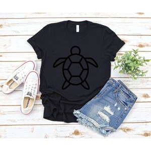 Puede incluir: Camiseta negra con un dise&ntilde;o de contorno de tortuga. La camiseta se combina con pantalones cortos de mezclilla y zapatillas blancas. Los art&iacute;culos est&aacute;n dispuestos sobre una superficie de madera blanca, creando una est&eacute;tica casual y veraniega.