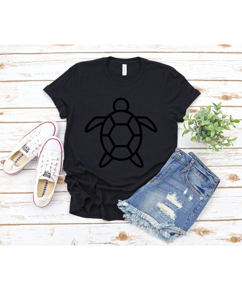 Puede incluir: Camiseta negra con un dise&ntilde;o de contorno de tortuga. La camiseta se combina con pantalones cortos de mezclilla y zapatillas blancas. Los art&iacute;culos est&aacute;n dispuestos sobre una superficie de madera blanca, creando una est&eacute;tica casual y veraniega.