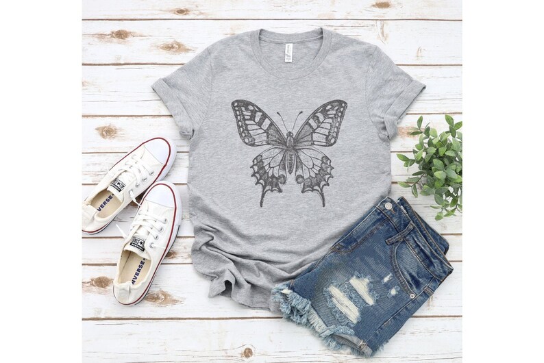 May include: A gray t-shirt with a black and white butterfly graphic. The shirt is paired with denim shorts and white sneakers.