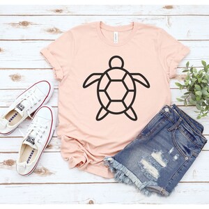 Puede incluir: Una camiseta rosa claro con un contorno negro de tortuga. Se combina con pantalones cortos de mezclilla y zapatillas de lona blancas. Los art&iacute;culos est&aacute;n colocados sobre una superficie de madera clara.