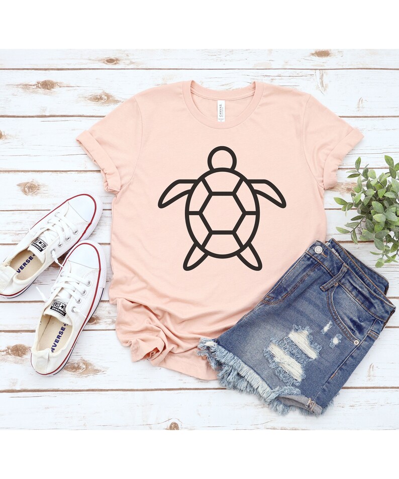 Puede incluir: Una camiseta rosa claro con un contorno negro de tortuga. Se combina con pantalones cortos de mezclilla y zapatillas de lona blancas. Los art&iacute;culos est&aacute;n colocados sobre una superficie de madera clara.