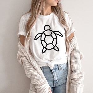 Puede incluir: Camiseta blanca con un contorno negro de una tortuga. El dise&ntilde;o de la tortuga est&aacute; centrado en la camiseta. La camiseta se lleva con vaqueros azules y una rebeca beige. La camiseta es de un material suave.