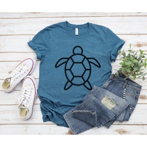 Puede incluir: Una camiseta de color turquesa con un gr&aacute;fico de tortuga negra, combinada con vaqueros de mezclilla y zapatillas de lona blancas. La camiseta presenta un dise&ntilde;o de tortuga geom&eacute;trico simple. El atuendo informal est&aacute; dispuesto sobre una superficie de madera clara.