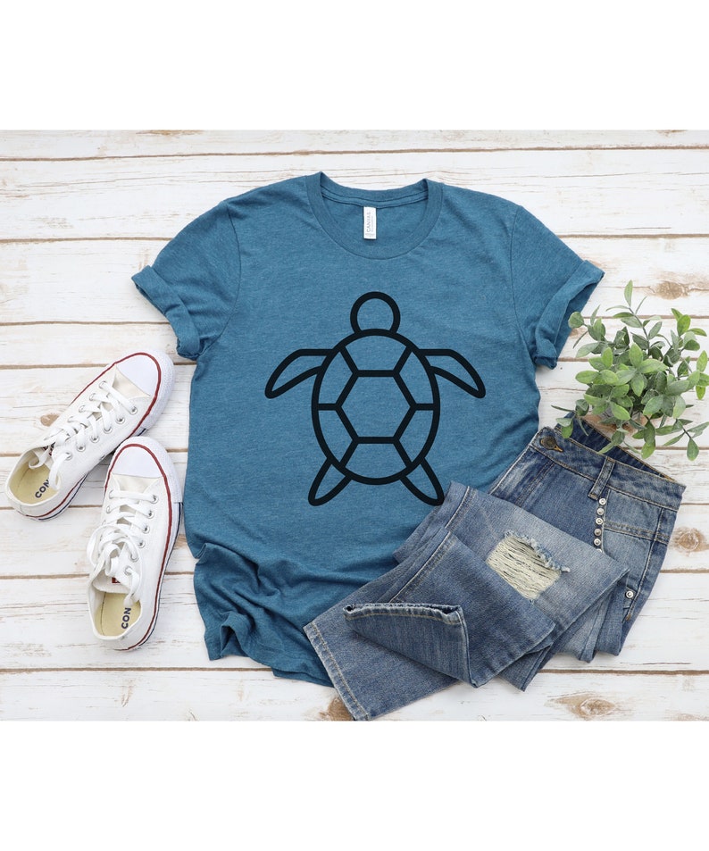 Puede incluir: Una camiseta de color turquesa con un gr&aacute;fico de tortuga negra, combinada con vaqueros de mezclilla y zapatillas de lona blancas. La camiseta presenta un dise&ntilde;o de tortuga geom&eacute;trico simple. El atuendo informal est&aacute; dispuesto sobre una superficie de madera clara.