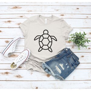 Puede incluir: Una camiseta de color beige claro con un dise&ntilde;o de tortuga negra. La camiseta se combina con pantalones cortos de mezclilla y zapatillas de lona blancas. Una peque&ntilde;a planta en maceta se encuentra cerca sobre una superficie clara con vetas de madera.
