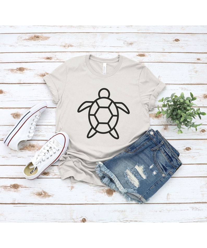 Puede incluir: Una camiseta de color beige claro con un dise&ntilde;o de tortuga negra. La camiseta se combina con pantalones cortos de mezclilla y zapatillas de lona blancas. Una peque&ntilde;a planta en maceta se encuentra cerca sobre una superficie clara con vetas de madera.