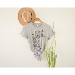May include: A gray t-shirt with a black butterfly graphic design. The butterflies are arranged in a cascading pattern on the front of the shirt.