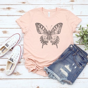 May include: A light pink t-shirt with a black and white butterfly graphic print. The t-shirt is paired with distressed denim shorts and white sneakers.
