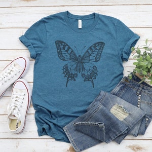May include: A teal blue t-shirt with a black and white butterfly graphic print. The shirt is laid out on a white wooden surface with a pair of white sneakers and a small potted plant.
