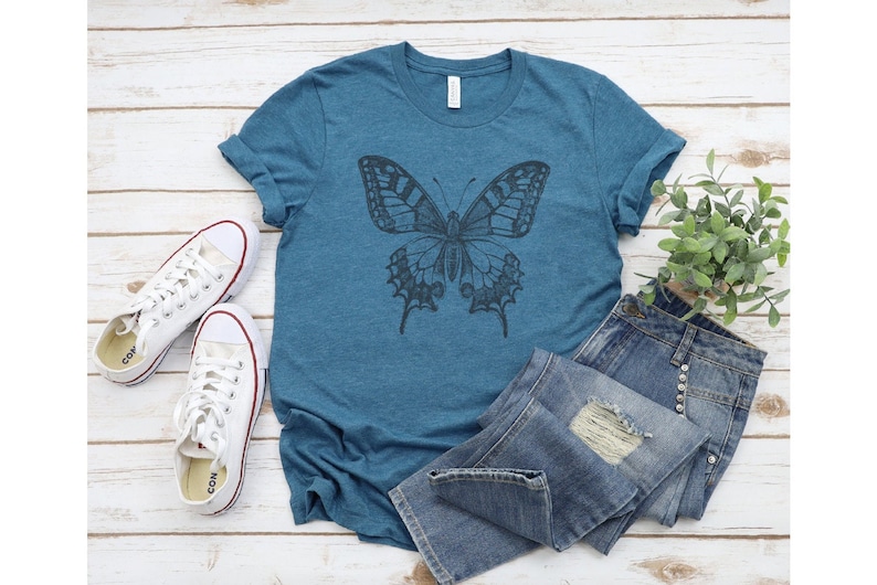 May include: A teal blue t-shirt with a black and white butterfly graphic print. The shirt is laid out on a white wooden surface with a pair of white sneakers and a small potted plant.