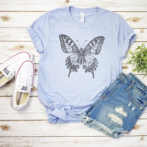 May include: A light blue t-shirt with a black and white butterfly graphic. The shirt is paired with denim shorts and white sneakers.