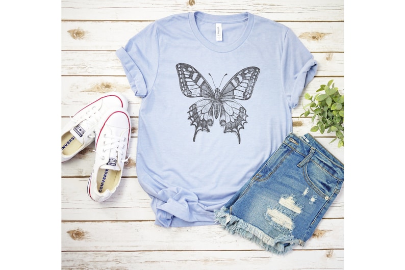 May include: A light blue t-shirt with a black and white butterfly graphic. The shirt is paired with denim shorts and white sneakers.