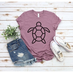 Puede incluir: Camiseta malva con un dise&ntilde;o de tortuga negra. La camiseta se combina con pantalones cortos de mezclilla y zapatillas de lona blancas. El conjunto se presenta sobre una superficie clara con efecto de madera, creando un ambiente veraniego relajado.