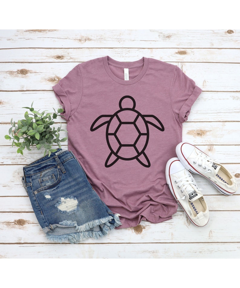 Puede incluir: Camiseta malva con un dise&ntilde;o de tortuga negra. La camiseta se combina con pantalones cortos de mezclilla y zapatillas de lona blancas. El conjunto se presenta sobre una superficie clara con efecto de madera, creando un ambiente veraniego relajado.