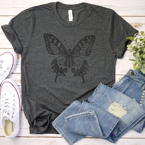 May include: A dark gray t-shirt with a black butterfly graphic design. The shirt is laid flat on a white wooden surface with a pair of white sneakers and blue jeans.