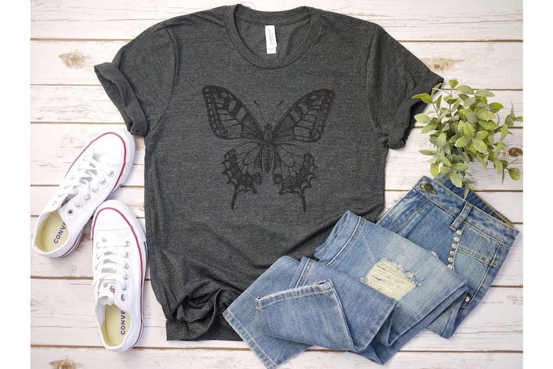 May include: A dark gray t-shirt with a black butterfly graphic design. The shirt is laid flat on a white wooden surface with a pair of white sneakers and blue jeans.