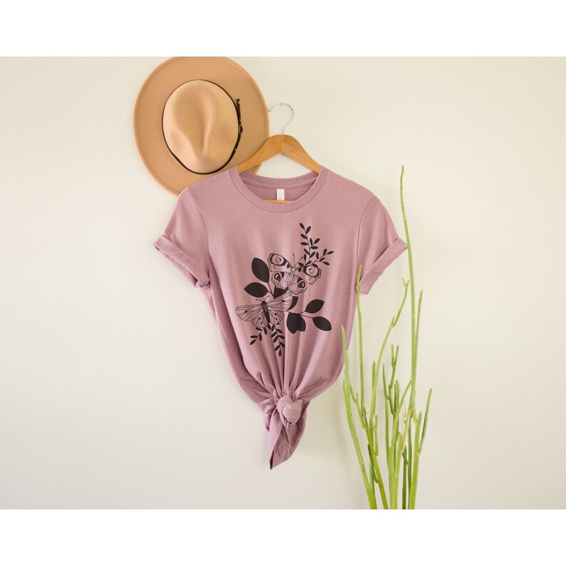 May include: A mauve t-shirt with a black graphic of a butterfly and flowers. The t-shirt is tied at the bottom and hanging on a hanger.