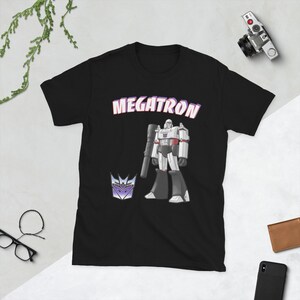 May include: Black t-shirt with a white and gray image of Megatron, a robot character from the Transformers franchise. The text "Megatron" is printed in pink above the image. A purple and gray symbol is printed in the bottom left corner.