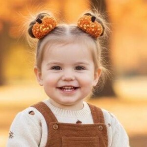 May include: A smiling toddler with pumpkin-shaped hair accessories. She wears a cream-coloured sweater with acorn embroidery and brown corduroy dungarees. The background is a blurred autumn scene.