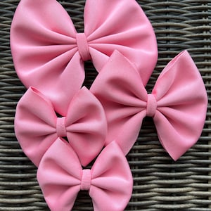 May include: Four pink fabric hair bows of different sizes. The bows are arranged in a pyramid shape on a woven brown background.