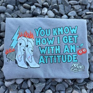 May include: Gray t-shirt with the text "You Know How I Get With An Attitude." The design includes illustrations of cowboy boots with flames, a lightning bolt, playing cards, cherries, and money.