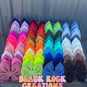 May include: A collection of 36 colorful hair bows arranged in a grid pattern. The bows are made of a soft, textured fabric and are tied with a double knot. The bows are displayed on a black metal tray with the text "BLACK ROCK CREATIONS" in pink and white letters.