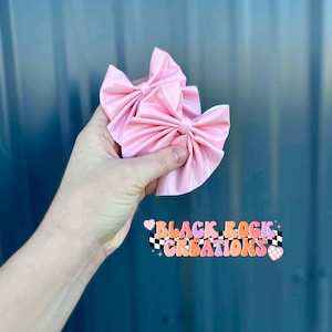 May include: Two light pink satin hair bows. The bows are held in a hand. The text "BLACK ROCK CREATIONS" is displayed in a colorful, retro font with a checkered pattern.