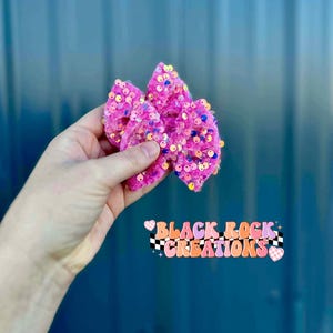 May include: A bright pink hair bow with sequins. The bow is held in a hand against a blue background. The bow is made of a soft, textured material and is covered in colorful sequins. The text "BLACK ROCK CREATIONS" is below the bow.
