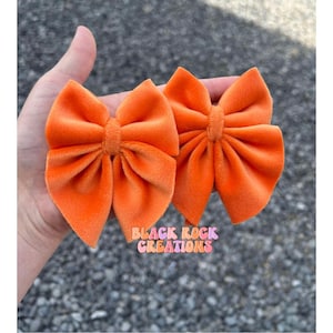 May include: Two orange velvet hair bows. The bows are tied in a simple knot and have a soft, plush texture.