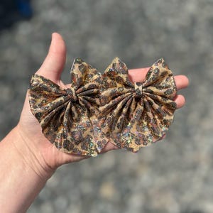 May include: Two leopard print hair bows with a glittery finish. The bows are held in a hand, showcasing their detailed pattern and texture. The bows are a mix of brown, tan, and black, with a shiny, iridescent effect.