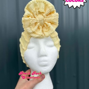 May include: A pale yellow turban-style head wrap with a large, ruffled bow on top. The head covering is displayed on a white mannequin head. The text "MORE COLORS" is in a pink starburst. The text "Black Rock Creations" is in pink script.