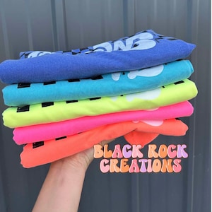 May include: Stack of five t-shirts in various bright colors, including neon green, pink, and orange. The shirts are folded and stacked on top of each other. The text "Black Rock Creations" is visible in a colorful font at the bottom of the image.