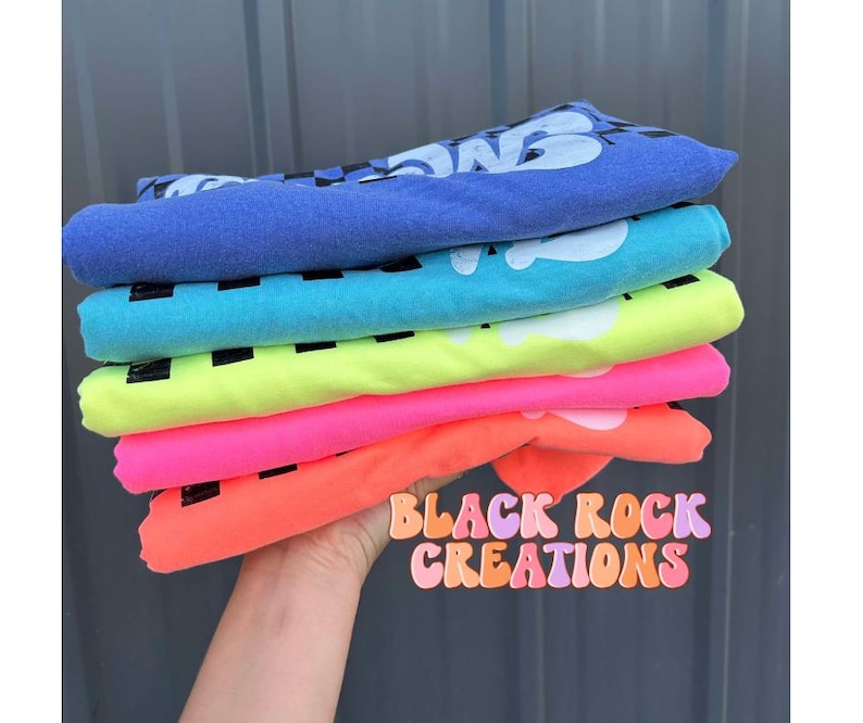 May include: Stack of five t-shirts in various bright colors, including neon green, pink, and orange. The shirts are folded and stacked on top of each other. The text "Black Rock Creations" is visible in a colorful font at the bottom of the image.