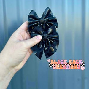 May include: Two black hair bows with a glossy leopard print pattern. The bows are tied in the center with a matching fabric. The bows are held in a hand. The text "BLACK ROCK CREATIONS" is in the bottom right corner.