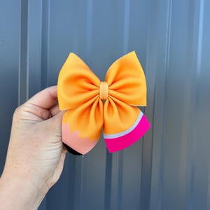 May include: A bright orange hair bow designed to resemble a pencil. The bow features a pink and gray eraser end, a black pencil tip, and a yellow-orange bow. This accessory is perfect for back-to-school or everyday wear.