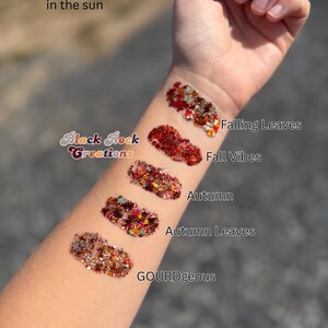 May include: Five distinct swatches of colorful glitter applied to an arm, labeled with the text Black Rock Creations, Photo taken in the sun, Falling Leaves, Fall Vibes, Autumn, Autumn Leaves, and GOURDgeous. The glitter features red, orange, gold, and brown tones.
