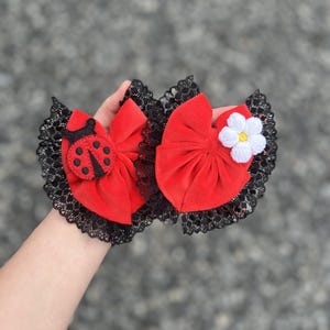May include: Two red velvet hair bows with black lace trim. One bow has a ladybug applique and the other has a white crocheted flower.