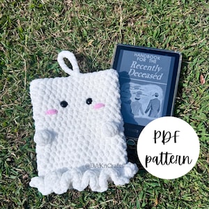 May include: White crocheted ghost amigurumi pattern with pink cheeks and black eyes. The pattern is available as a PDF download. The ghost is sitting on green grass next to a book titled "Handbook for the Recently Deceased".