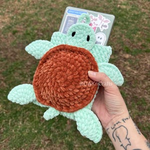 May include: A crocheted turtle toy with a green shell and brown head. The turtle is held in a hand with a tattoo that says "I love you".