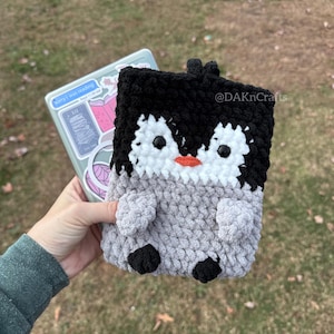 May include: A crocheted penguin phone case in black, white, and gray with a black nose and orange beak. The case is being held in a hand with a sticker on the phone that says "sorry I was reading".