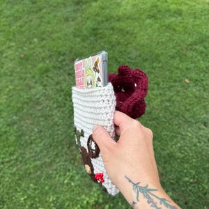 May include: A crocheted phone case in cream, brown, and burgundy. The case features a house design with a door and window, and a burgundy pouch. A phone with a clear case and a cartoon graphic is inside the case.