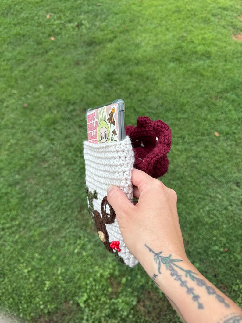 May include: A crocheted phone case in cream, brown, and burgundy. The case features a house design with a door and window, and a burgundy pouch. A phone with a clear case and a cartoon graphic is inside the case.