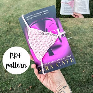 Puede incluir: Marcapáginas de ganchillo rosa en forma de triángulo. El marcapáginas está colocado en un libro titulado "The Love Hypothesis" de Ali Hazelwood. El libro está sostenido por una mano con un tatuaje de un elefante.
