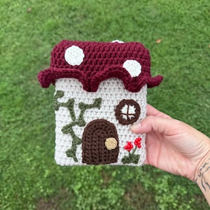 Puede incluir: Una casa de setas hecha a mano con ganchillo, con un tejado burdeos con lunares blancos. La casa tiene un cuerpo color crema, una puerta marrón, una ventana redonda y enredaderas verdes. Flores rojas añaden un toque decorativo.