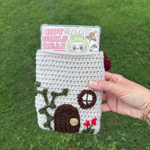 May include: A crocheted phone case in the shape of a house, with a brown door, green vines, and red flowers. The case holds a phone with a sticker that says "HOT GIRLS READ". The case is held against a green grass background.